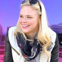 Allie Oosta, wearing a leather jacket and sunglasses, smiles at something off-camera.